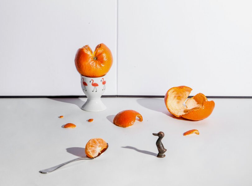 A half peeled tangerine onto of an egg cup, peelings teaspoon and small metal duck figurine spread onto table top