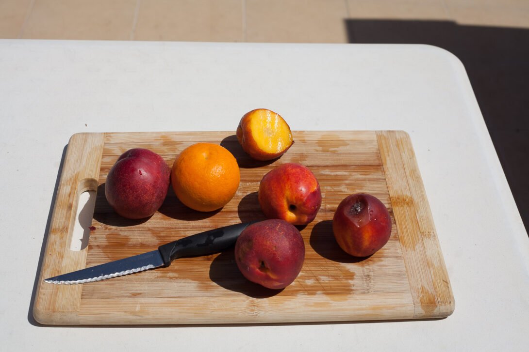 A bunch of wet peaches and one orange on a wooden chopping board with knife