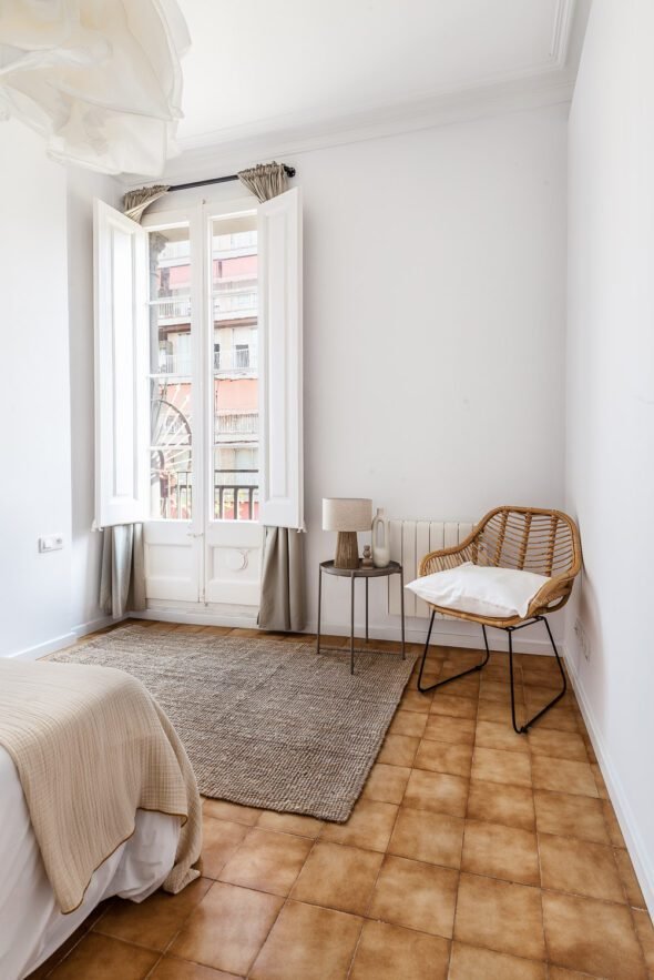 Single bedroom with a wicker chair, side table, lamp and french style doors with view outside