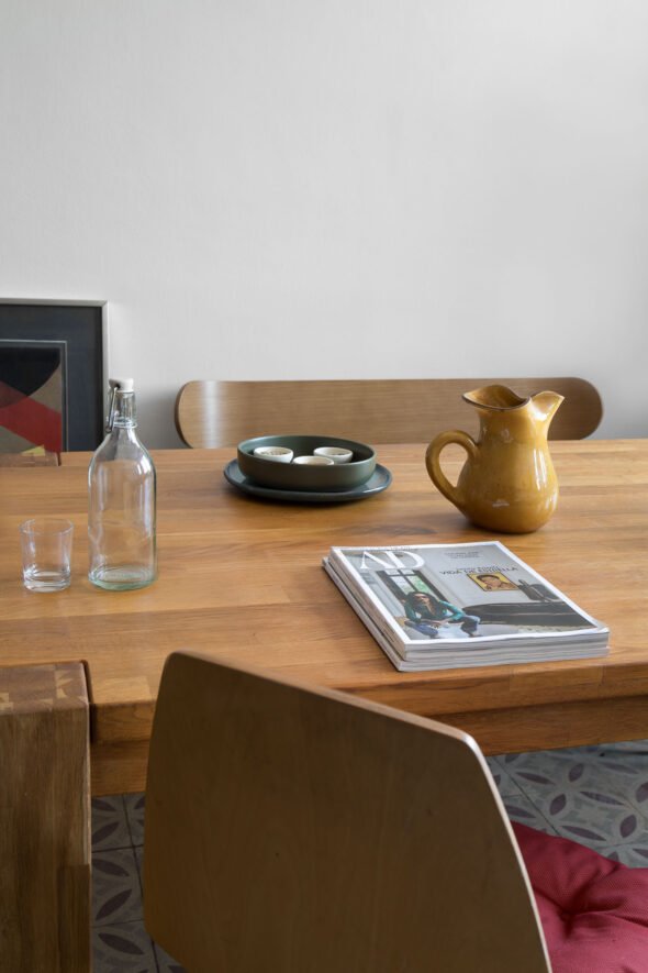 Still life interior photography featuring curated objects such as a yellow ceramic curly lipped vase dark green bowl, glass bottle and a selection of AD magazines styled on top of a wooden table.