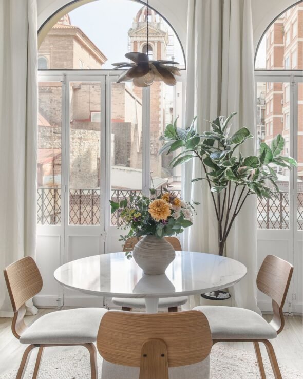 a small round marble dingtable with flowers underneath a bronze metal lampshade and a view out of large arched catalan window into court yard in barcelona
