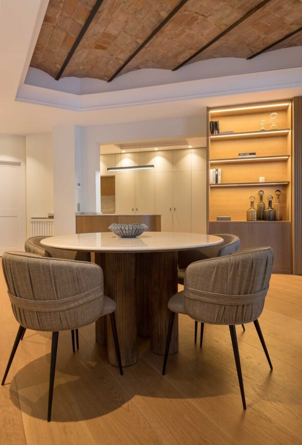 integrated dining area, bespoke bookshelf, catalan ceiling, contemporary kitchen in background, warmly lit ambient space.