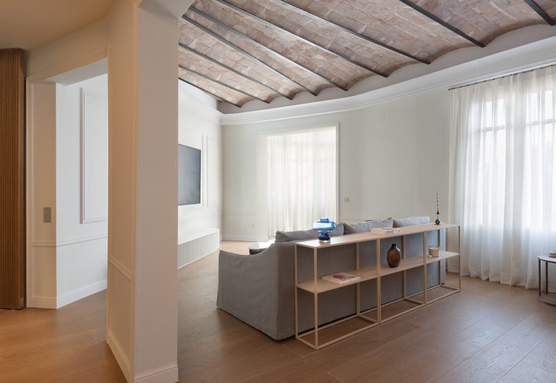 contemporary living room in barcelona eixample, beautiful diffused natural light, slanted catalan ceiling, white linen curtains, sofa, shelf with objects and white pilar in barcelona