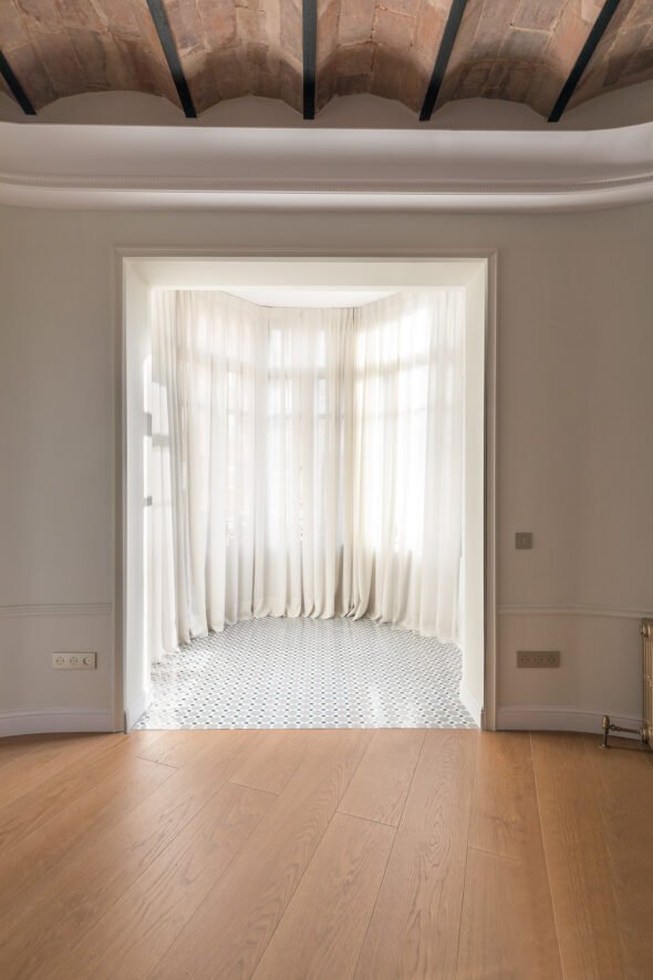 a symmetrical image of a view into a empty room with beautiful diffused natural light and white linen curtains, from an empty living room.wood floor and catalan ceiling.